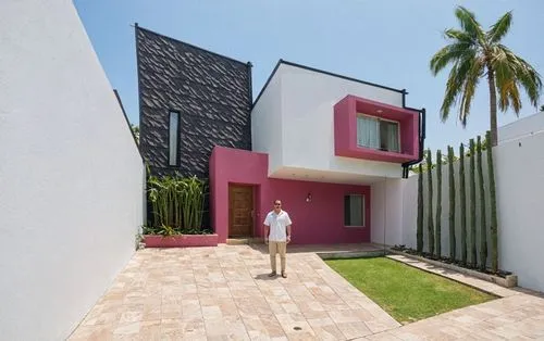 A man standing in the middle of the image, wearing a white shirt and khaki pants, in front of a modern, minimalist house with a pink and white exterior and a black shingled roof. the house has a recta