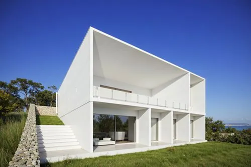 A photo-realistic shoot from the side about a modern, minimalist white house with a triangular roof and large glass windows, set against a clear blue sky. the house is situated on a hillside, with a g