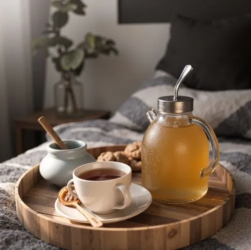 A wooden tray with a glass teapot, a cup of tea, honey, and cookies placed on a plush gray bed, capturing a serene and inviting morning setup.,a glass pitcher on top of a wooden tray next to a cup,tea