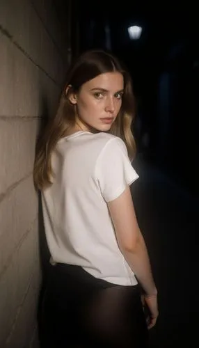 A woman around 20s, long brown hair (tips of hair being light brown) and dark eyes, in a white t-shirt and black tights,  standing in a dark alley standing while back turned on a wall, with light comi