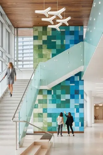 A photo-realistic rendering of a modern, high-tech building with a large, colorful, geometric wall featuring green and blue tiles. The chandeliers are on. The image also shows a staircase with a glass
