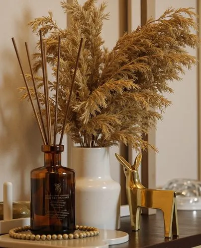 An elegant arrangement of home decor items, including a golden deer figurine, a dark amber reed diffuser, and dried wheat in a glass vase. The setting is enhanced with delicate lighting and soft textu