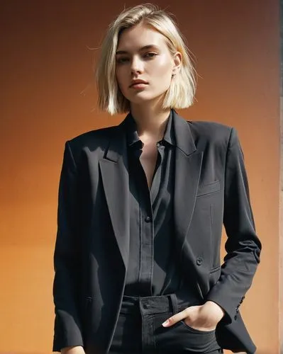 An edgy model with blond medium-length hair. She wears a black oversize blazer outfit. The model stands cool, straight, and relaxed in front of the camera. It's a studio shot. The background has a gra