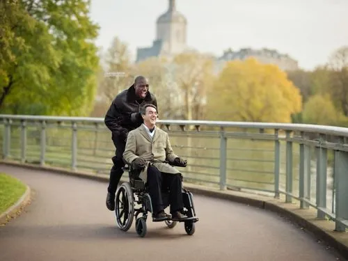 A man pushing another man in a wheelchair, on a bridge in a park in the background,the older man is riding an unicycle with his companion,elderly couple,velib,care for the elderly,caregiving,handcycle