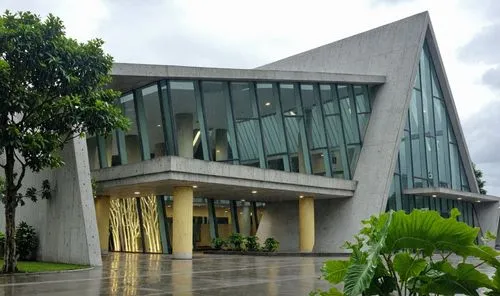 A photo of a building. The light gray concrete and large areas of glass make up its facade. The light yellow pillars support the building. There are a large number of plants surrounding it. The whole 