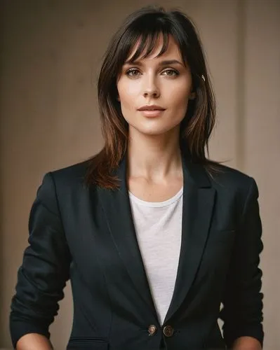 A sassy model with straight, long, dark hair. She is wearing a black, loose blazer. The model stands straight in front of the camera, calm and relaxed. This is a studio shot. The background has a grad