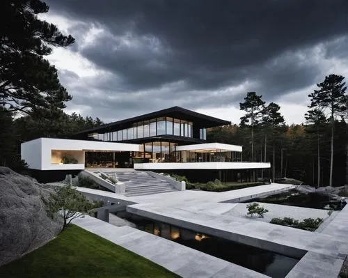 A striking modern house perched on a cliff, featuring sharp geometric shapes and floor-to-ceiling glass windows. The home is surrounded by dense pine trees, with dramatic lighting emphasizing its futu