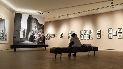 the sitting woman is looking at a black and white photograph in a museum. in the photograph, a man is posing with some ancient ruins,man sitting on bench near wall with art,fotomuseum,gallery,exhibiti