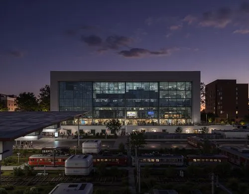 A photo-realistic shoot from a bird's eye view about a large, modern building lit up at night, with multiple train cars parked at a train station in the foreground. the image also shows a long exposur