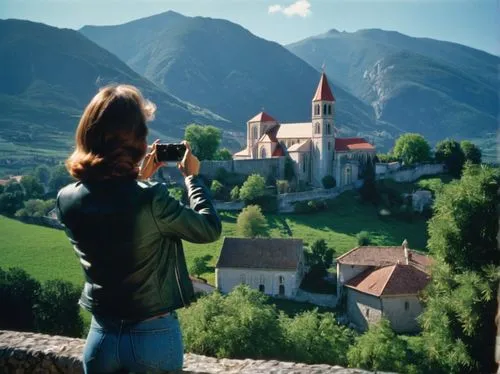 A woman takes pictures of the monastery at Crie in France, surrounded by mountains and blue sky, while wearing jeans with a leather jacket, looking over stone walls to see a church with a red roof. Th
