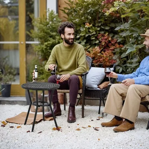 On the white gravel floor, next to a couple sitting in chairs drinking wine. In the background are plants in the garden,two men sitting on chairs outside with bottles of wine in their hands,echenique,