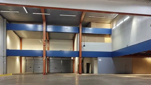 “Vista interior de una bodega industrial moderna, caracterizada por amplios espacios abiertos, estructuras metálicas en color negro y columnas de madera que soportan entrepisos. Las paredes en tonos g