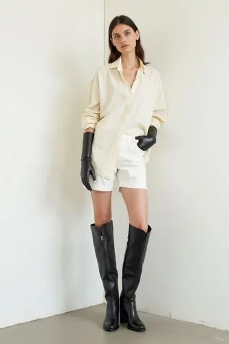 She stands naturally in front of a white wall. She wears a cream oversized button-down shirt tucked loosely into a pair of white cotton tailored shorts. Over the arms, glossy black leather gloves exte
