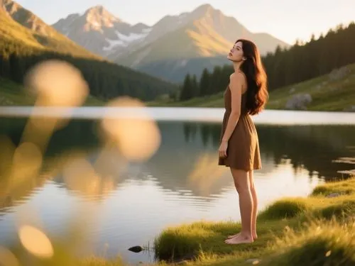 A realistic outdoor portrait photograph captured during golden hour at a serene alpine lake, featuring a young woman with warm golden-tanned skin and long flowing dark brown hair standing barefoot at 