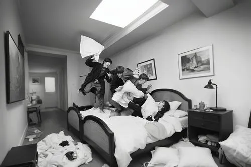 A wider black-and-white view of the room, revealing the chaos and laughter spilling beyond the bed. The floor is scattered with pillows and blankets, some halfway off the bed. A small bedside table ho
