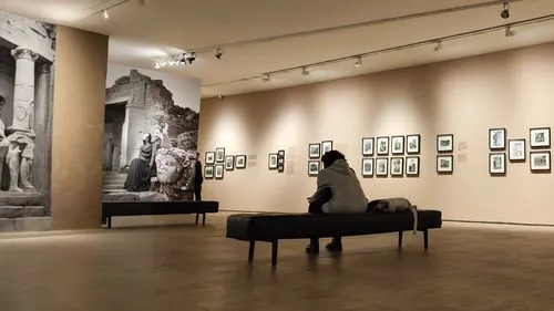 the sitting woman is looking at a black and white photograph in a museum. in the photograph, a man is posing with some ancient ruins,there is a woman sitting on a bench in the middle of a museum,galle