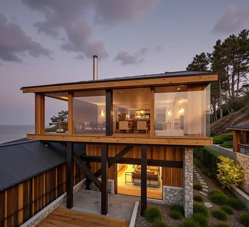 A photo-realistic shoot from a bird's eye view about a modern house with a wooden exterior and glass walls, situated on a hillside overlooking the ocean at dusk. the house has a black metal roof and a