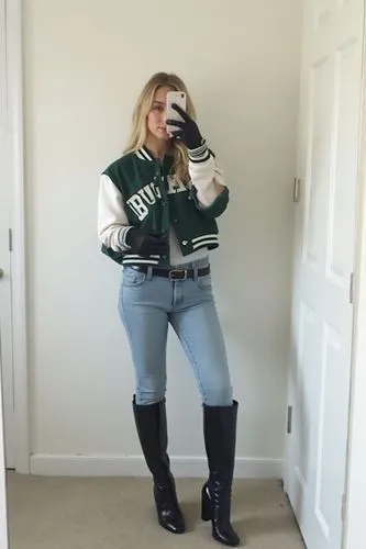 She’s in a basic white room, standing with one leg bent, phone blocking half her face. She wears a cropped graphic varsity jacket in forest green and white, sleeves cut short to expose black leather g