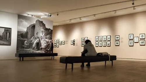 the sitting woman is looking at a black and white photograph in a museum. in the photograph, a man is posing with some ancient ruins,a person sitting on a bench in front of pos,fotomuseum,gallery,exhi