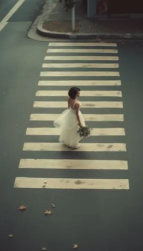 A high-angle shot of a lone person walking across a wide crosswalk, similar to a zebra crossing, with strong horizontal lines dividing the road. The person is dressed in wedding dress holding bouquet 