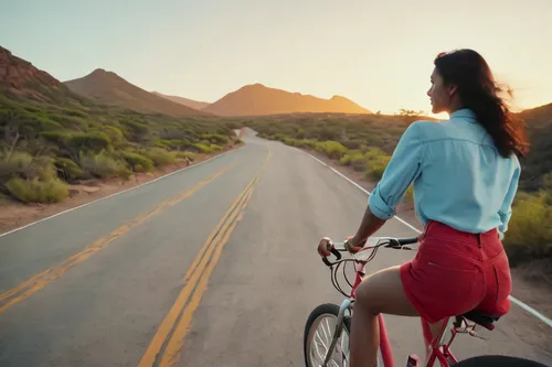 Photograph taken from a ground angle and from behind of a woman with wavy black hair and white skin, dressed in very short jean shorts, a light blue long-sleeved shirt, and red Converse shoes, riding 