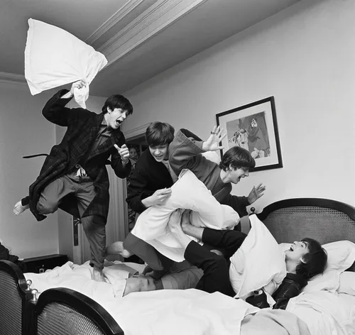 A lively black-and-white photograph capturing a playful moment of four young men engaged in a spirited pillow fight on a large, disheveled bed. Each figure is full of energy: one mid-leap with an exag