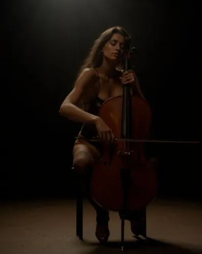 A photograph of a beautiful young caucasian woman with long brown hair sitting on a stool with a cello, playing the instrument on a dimly lit stage in a dark theater. She is wearing a black swimsuit a