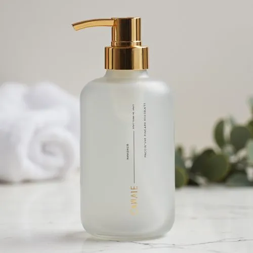 A sleek cylindrical personal care bottle made of frosted glass with a smooth matte finish, standing upright on a white marble surface. The bottle features a minimalist label with clean black sans-seri