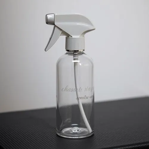 A sleek spray bottle placed on a matte black surface with a soft spotlight emphasizing its clean, contemporary design. The bottle is made of frosted glass with a cylindrical shape, featuring a minimal