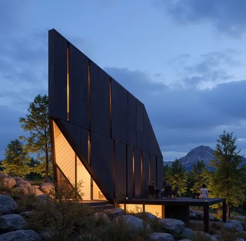 A striking modern black house with angular architecture, featuring floor-to-ceiling windows and a sharp geometric design. The house is set in a rocky landscape surrounded by trees, blending dramatic m