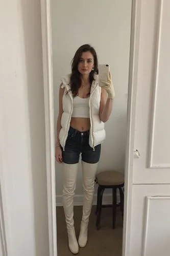 The selfie is spontaneous and unposed—mirror is slightly dirty, phone flash is on. She’s standing loosely, one hand on her hip, not even looking directly at the camera. White walls and a corner of a c