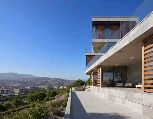 A photograph taken from a high vantage point, looking down on a modern building with large windows and a glass railing. the building is situated on a hillside, with a clear blue sky in the background.