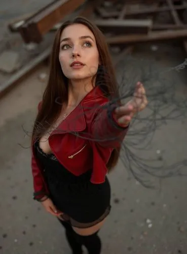 A photograph of a beautiful caucasian young woman, staning in a street. She has long brown hair, is wearing a red leather jacket and a black minidress whit a neckline and longblack loots . She has her