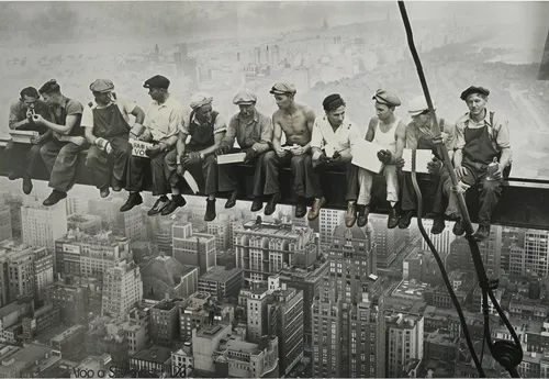 A black-and-white photograph of eleven construction workers sitting casually on a suspended steel beam high above a bustling 1930s New York City skyline. The workers, dressed in rugged work attire wit