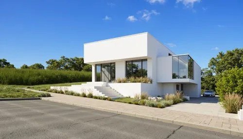 A modern, minimalist white house with large glass windows and a grassy hillside. the house is situated on a serene, suburban street, with a clear blue sky and a few scattered clouds in the background.