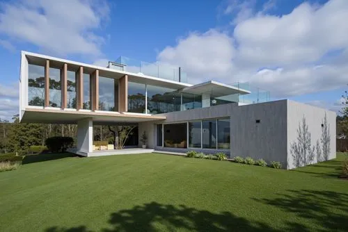 A photo-realistic shoot from a bird's eye view about a modern concrete house with large glass windows on a hillside, set against a bright blue sky with fluffy white clouds. the house is situated on a 