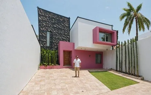A man standing in the middle of the image, wearing a white shirt and khaki pants, in front of a modern, minimalist house with a pink and white exterior and a black shingled roof. the house has a recta