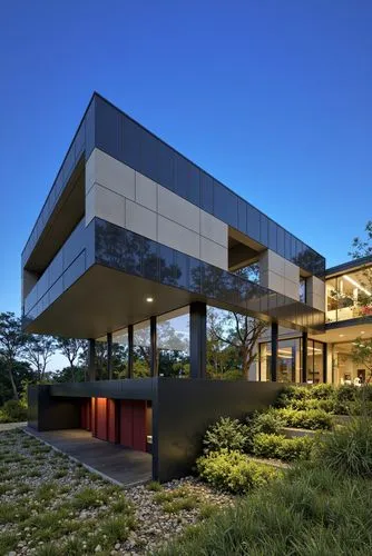 
A modern, minimalist house with a dark grey exterior and large glass windows, situated on a hillside with a clear blue sky in the background. the house is situated on the right side of the image, wit
