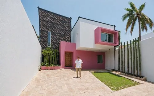 A man standing in the middle of the image, wearing a white shirt and khaki pants, in front of a modern, minimalist house with a pink and white exterior and a black shingled roof. the house has a recta