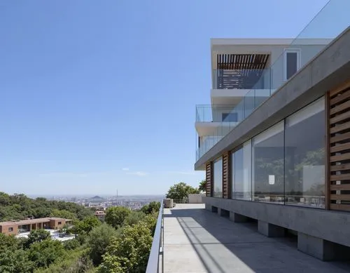 A photograph taken from a high vantage point, looking down on a modern building with large windows and a glass railing. the building is situated on a hillside, with a clear blue sky in the background.
