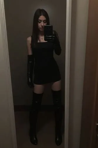 A dark, low-light mirror selfie taken quickly with a phone, slightly blurry and grainy as if shot in a dim bedroom or hallway. An 18-year-old woman poses casually as if it were a normal outfit-of-the-
