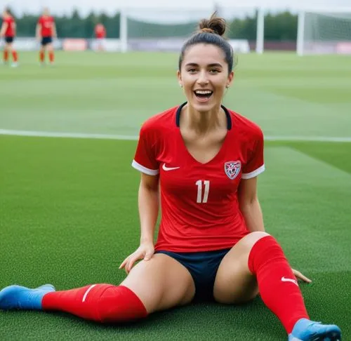 Same person wearing soccer cleats, soccer socks, soccer pants and a bra. She scored a goal on a soccer pitch. She celebrates by knee sliding and holding her red jersey in her hand. She is wearing a br