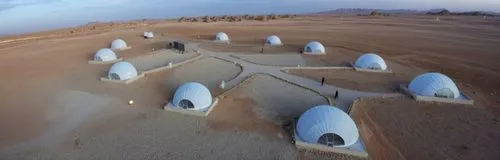 The camp in the desert of KSA contains a group of white geodesic dome tents, and there are lots of landscape lights to define the road between the tents. Every tent has beside it an Arabic seat and a 