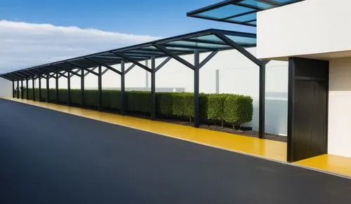 A clean and modern outdoor walkway with a series of metal posts supporting a glass roof. The posts are matte black. The setting features well-manicured hedges on the side, a smooth road with yellow li