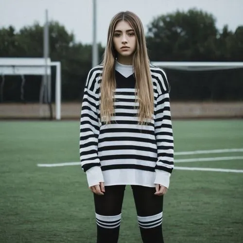 A young 20 year old girl with long, straight light brown hair is standing on a soccer field which has a water on it. She is wearing a black and white striped long-sleeve sweater and knee-high black so