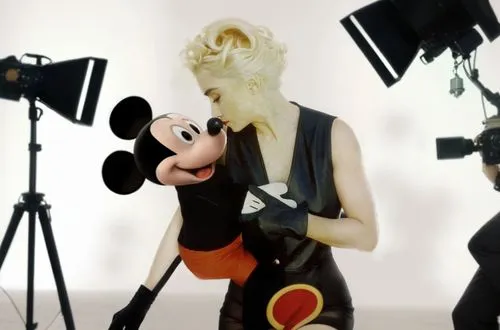 Madonna is on a movie set with Mickey Mouse in her arms!  Madonna is wearing a sexy dress and has beautiful legs. The movie set is full of crew members such as: director, camera operator, sound contro