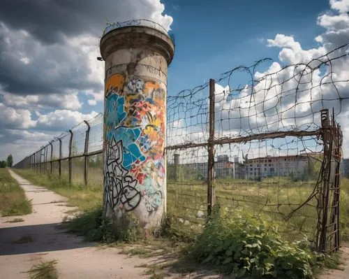 Berlin Wall, historic landmark, graffiti-covered concrete barrier, broken remains, urban decay, abandoned, overgrown with weeds, rusty barbed wire, surveillance tower, somber mood, cloudy sky, afterno