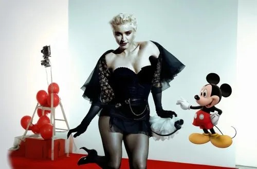 Madonna is on a movie set with Mickey Mouse in her arms!  Madonna is wearing a sexy dress and has beautiful legs. The movie set is full of crew members such as: director, camera operator, sound contro