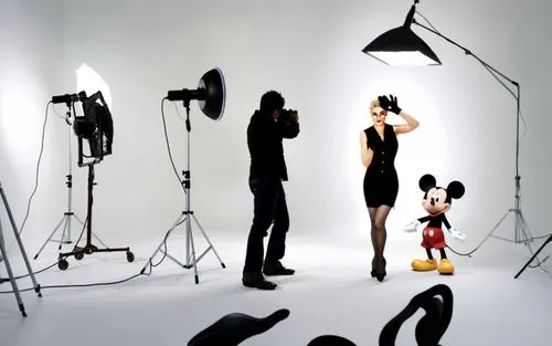 Madonna poses for fashion photos with Mickey Mouse. The photographer shoots the scene with other crew members in a studio. Madonna wears a short skirt where she shows off her legs with dark stockings 