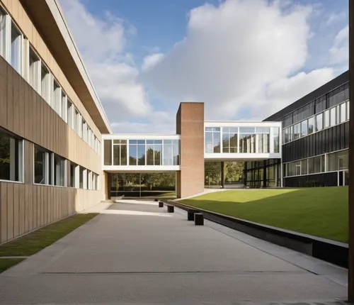 a building with straight lines, with colored panels on the facade, windows with dark frames, in the background a glass passage with a concrete pillar in the center and trees behind, a garden on the ri